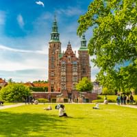 Rosenborg Castle with red brick towers and green copper spires in Copenhagen, surrounded by park lawns with visitors relaxing under blue sky.