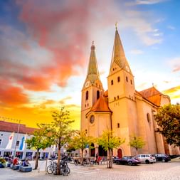 St. Walburga Kirche mit zwei Türmen am Marktplatz in Beilngries bei Sonnenuntergang. Farbenfroher Himmel, umliegende Gebäude, Fahrräder und Autos.