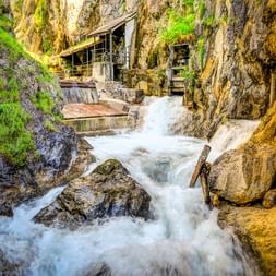 Tossendes Wasser fließt durch die enge Lötzklamm in Österreich. Moosbewachsene Felswände rahmen den Bach, darüber sind Holzstege sichtbar.