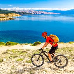 Radfahrer in rotem Trikot mit orangem Rucksack fährt Mountainbike auf Küstenweg in Kroatien mit blauem Meer und Bergen im Hintergrund.