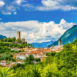 Gemona del Friuli eingebettet in grüne Hügel mit mittelalterlichem Turm auf Hügel, Kirchturm und dramatischer Bergkulisse unter blauem Himmel.