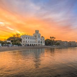 Weiße Festung Neratzia an der Uferpromenade von Kos bei Sonnenuntergang mit orangefarbenem Himmel, Palmen und ruhigem Meer.