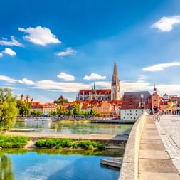 Historische Steinerne Brücke über die Donau zur Regensburger Altstadt mit Domturm und bunten mittelalterlichen Gebäuden unter blauem Himmel.
