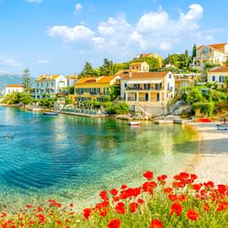 Fiscardo-Bucht mit bunten mediterranen Häusern am Ufer, roten Mohnblumen im Vordergrund, kleinem Strand, Booten im klaren Wasser und Bergen.
