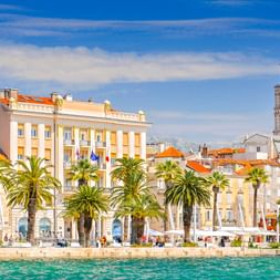 Split Uferpromenade mit Palmen, historischen Gebäuden mit orangen Dächern und dem Glockenturm der Kathedrale unter blauem Himmel.