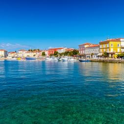 Hafen von Poreč mit bunten historischen Gebäuden entlang der Uferpromenade, Boote im klaren türkisfarbenen Wasser unter blauem Himmel.