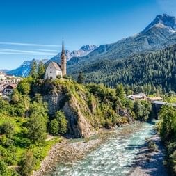 Weiße Kirche mit hohem Turm auf Felsvorsprung über türkisfarbenem Fluss, umgeben von Alpendorf, Wäldern und dramatischen Berggipfeln.