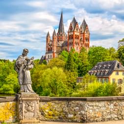 Steinstatue auf Brücke mit Blick auf den Limburger Dom mit sieben Türmen über grünen Bäumen und gelbem Wohnhaus unter bewölktem Himmel.