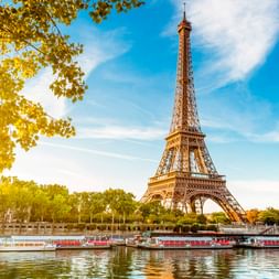 Der Eiffelturm erhebt sich über der Seine in Paris an einem sonnigen Tag. Ausflugsboote liegen am Flussufer, eingerahmt von grünen Bäumen.