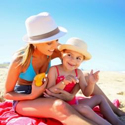 Frau und kleines Mädchen in Badeanzügen und Sonnenhüten sitzen auf rotem Handtuch am Sandstrand, lächeln zusammen unter blauem Himmel.