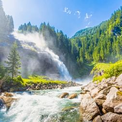 Krimmler Wasserfälle stürzen bewaldeten Berghang hinab mit Gischt, felsiger Bach im Vordergrund, umgeben von grünen Nadelwäldern.