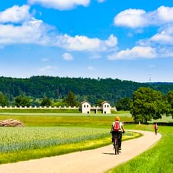 Zwei Radfahrer auf einem asphaltierten Weg durch grüne Felder zum rekonstruierten Römerkastell Pfünz, mit Wäldern und blauem Himmel im Hintergrund.