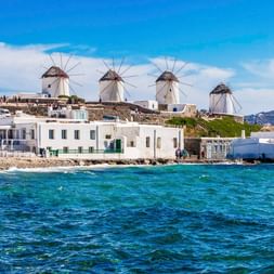 Fünf traditionelle weiße Windmühlen mit Blick auf das türkisfarbene Meer auf Mykonos. Weiße kubische Häuser säumen die Uferpromenade.