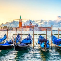 Reihe blauer Gondeln an Holzpfählen in Venedig mit der Kirche San Giorgio Maggiore über der Lagune unter dramatischem Abendhimmel.
