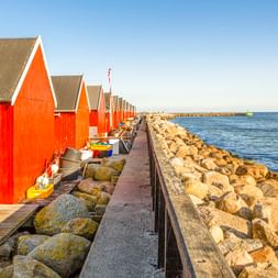 Reihe roter Holzbootshäuser mit weißen Verzierungen entlang eines Steinpiers im Hafen von Rødvig. Kleine Boote liegen neben den Gebäuden unter blauem Himmel.