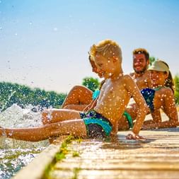 Drei Kinder sitzen auf einem Holzsteg und spritzen mit den Füßen Wasser. Wassertropfen glitzern im Sonnenlicht vor blauem Himmel und grünen Bäumen.