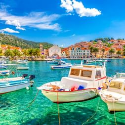 Hafen von Hvar mit weißen Booten in kristallklarem türkisfarbenem Wasser, Palmen, mediterranen Gebäuden und grünen Hügeln unter blauem Himmel.
