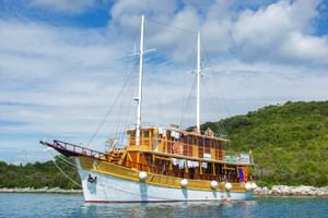 MS Planka, a wooden two-masted sailing ship with white hull and yellow-brown superstructure, anchored in calm blue water with green hills.