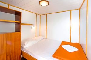 Compact cabin on MS Planka with double bed, white linens, orange towel, wooden storage, and white walls with wood trim.