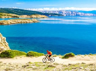 Cyclist in red jacket riding along coastal path overlooking turquoise Adriatic Sea with rocky cliffs and mountains in Croatia.