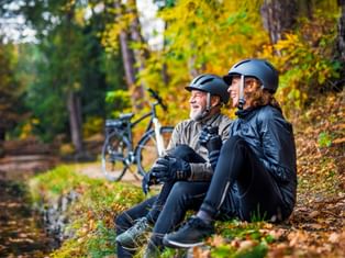 Two cyclists in helmets sitting by a forest stream in autumn, with e-bikes parked behind them. Colorful fall foliage surrounds the scene.