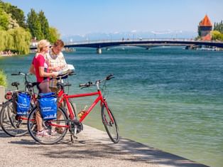 Zwei Radfahrer mit Rädern lesen eine Karte am Seerheinufer in Konstanz. Historischer Turm und Brücke über dem türkisfarbenen Wasser.