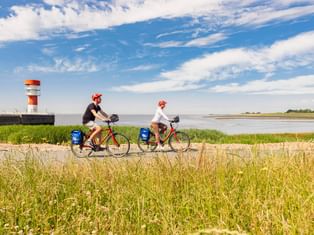 Zwei Radfahrer mit roten Helmen und blauen Taschen fahren am Uferweg in Brunsbüttel. Rot-weißer Leuchtturm und Elbe unter blauem Himmel sichtbar.