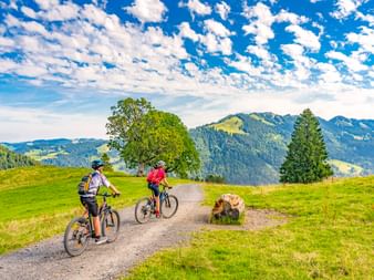 Zwei Radfahrer auf einem Schotterweg bei Oberstaufen mit grünen Wiesen, Bäumen und bewaldeten Bergen unter blauem Himmel mit weißen Wolken.
