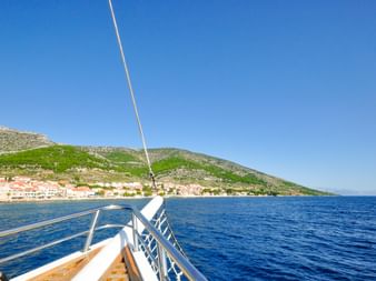 Blick vom Bootsdeck auf die Küste von Split mit Gebäuden am Ufer und grünen Hügeln unter klarem blauen Himmel. Tiefblaue Adria im Vordergrund.
