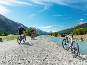 Zwei Radfahrer auf einem Schotterweg neben dem türkisfarbenen Inn in Österreich, mit Bergen und Wäldern im Hintergrund unter blauem Himmel.