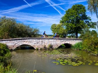 Zwei Radfahrer fahren über eine alte Steinbrücke über einen ruhigen Fluss mit Seerosen, umgeben von grünen Bäumen unter blauem Himmel.