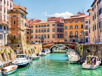 Historischer Kanal in Livorno mit vertäuten Booten, bunten Gebäuden und einer Steinbrücke. Klares türkisfarbenes Wasser spiegelt den Himmel.