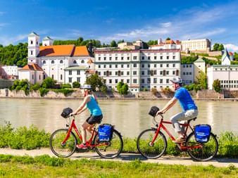 Zwei Radfahrer mit roten Rädern und blauen Taschen fahren am Fluss in Passau entlang, mit historischen Gebäuden und Kirche auf der anderen Seite.