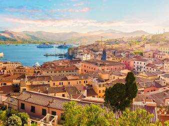 Luftaufnahme des Hafens von Portoferraio mit bunten Gebäuden, Terrakottadächern, Fähren in der Bucht und Bergen im Hintergrund bei Sonnenuntergang.
