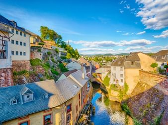Luftaufnahme der Saarburger Altstadt mit traditionellen Gebäuden entlang eines schmalen Kanals. Historische Häuser auf felsigem Hang unter blauem Himmel.
