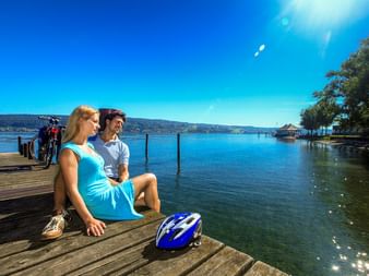 Zwei Radfahrer sitzen auf einem Holzsteg am Bodensee mit Fahrrädern hinter ihnen. Blauer Helm im Vordergrund, ruhiger See und sonniger Himmel.
