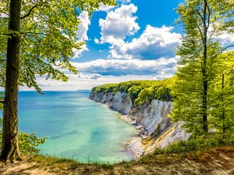 Weiße Kreidefelsen des Königsstuhls auf Rügen, eingerahmt von grünen Buchen, mit Blick auf die türkisfarbene Ostsee unter blauem Himmel mit Wolken.