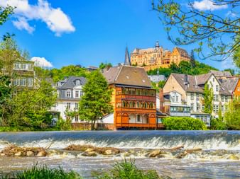 Marburger Landgrafenschloss auf Hügel über historischer Altstadt mit Fachwerkhäusern und Flusswehr im Vordergrund unter blauem Himmel.