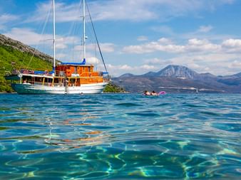 Segelschiff MS Mirabela aus Holz ankert in kristallklarem türkisfarbenem Wasser mit Bergen im Hintergrund. Schwimmer nahe dem Schiff sichtbar.