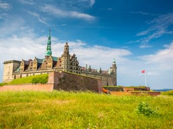 Schloss Kronborg in Helsingør mit Renaissance-Türmen und grünen Kupferspitzen, umgeben von grasbewachsenen Wällen. Dänische Flagge weht oben.