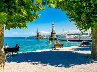 Blick durch Bäume auf den Konstanzer Hafen am Bodensee mit Imperia-Statue, Leuchtturm und Menschen auf Bänken am Ufer.