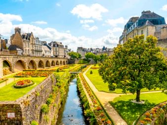 Formale Gärten mit bunten Blumenbeeten und Wasserkanal entlang alter Steinmauern und Bogenbrücke in Vannes, dahinter historische Gebäude.