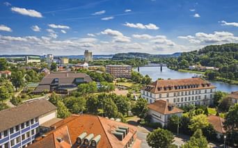 Luftaufnahme von Hameln mit rotgedeckten Gebäuden, der Weser, einer Brücke und grünen Hügeln unter blauem Himmel mit Wolken.