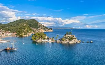 Zwei kleine Felseninseln mit grüner Vegetation in türkisfarbenem Wasser bei Parga. Ein Holzsegelboot ist sichtbar, mit Küstenstadt und Bergen im Hintergrund.
