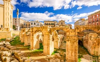 Ruinen des antiken römischen Amphitheaters in Lecce mit Steinsäulen und Bögen, umgeben von historischen Gebäuden unter blauem Himmel.