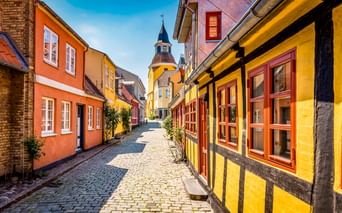 Kopfsteinpflasterstraße in Faaborgs Altstadt mit bunten Fachwerkhäusern in Gelb, Orange und Rot. Ein Kirchenglockenturm erhebt sich im Hintergrund.