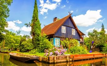 Blaues Holzhaus mit orangefarbenem Ziegeldach am Wasser in Lübben Spreewald. Holzsteg mit Blumen und Boot davor, umgeben von Bäumen.