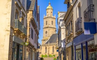Steinerner Glockenturm der Saint-Gildas-Kirche mit goldener Kuppel durch enge Gasse in Auray. Historische Gebäude säumen beide Seiten unter blauem Himmel.