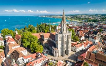 Luftaufnahme des Konstanzer Münsters mit gotischem Turm, umgeben von Gebäuden mit roten Dächern und dem Bodensee im Hintergrund.