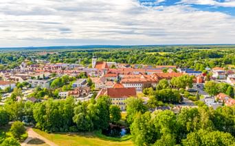 Luftaufnahme von Lübben mit roten Ziegeldächern, weißem Kirchturm, grünen Parks und Wäldern unter teilweise bewölktem Himmel.